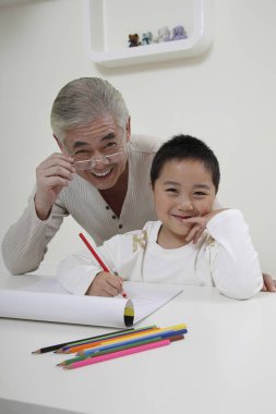 Çizim yaparken gülümseyen çocuk, arkasında duran kıdemli adam.