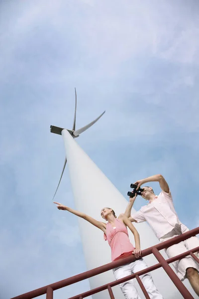 Woman pointing out over railing while man is looking through binoculars