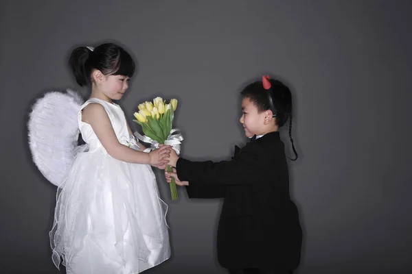 Boy with tuxedo and devil horns giving flowers to girl wearing white dress and angel wings