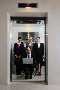 İş adamları ofis koltuğunda oturan ve asansörde dizüstü bilgisayar kullanan iş adamlarını izliyor.