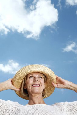 Yaşlı kadın şapkasını tutarken gülümsüyor.