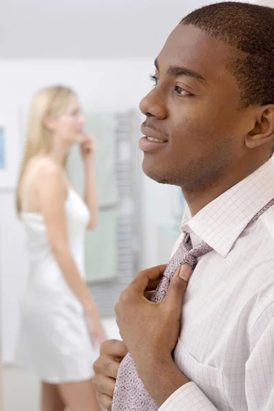 Man adjusting his necktie, woman applying blusher in the background