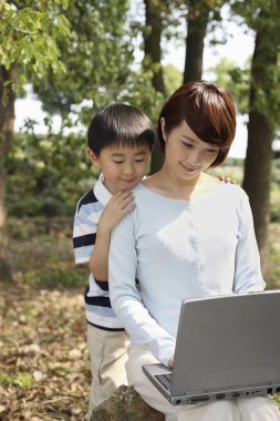 Kadın dizüstü bilgisayar kullanıyor, çocuk arkadan izliyor.