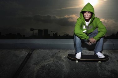 Yeşil kapüşonlu kazaklı adam kaldırımda oturuyor, kaykaya basıyor.