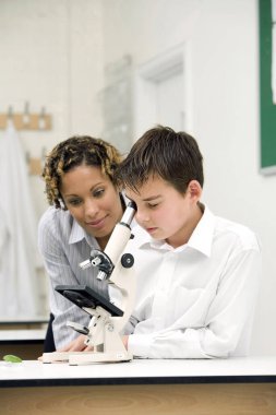 Kadın, mikroskopla bakan çocuğa yardım ediyor.