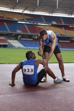 Pistte yatan erkek sporcu, bacağını acıdan kavrıyor, başka bir sporcu yardım ediyor.