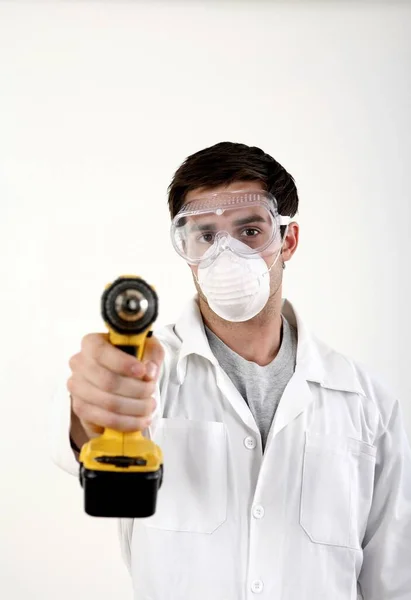 Man with safety mask and goggles pointing a power drill at the camera