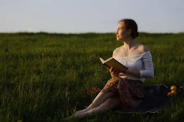 Jovencita leyendo un libro en la hierba mientras se pone el sol. Sensación de verano — Foto de Stock