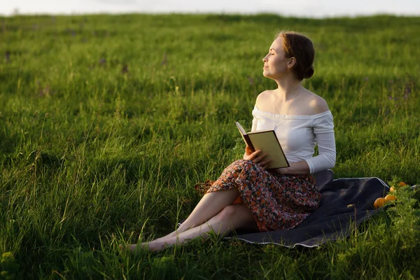 Jovencita leyendo un libro en la hierba mientras se pone el sol. Sensación de verano — Foto de Stock