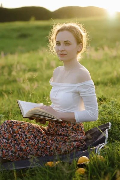 Jovencita leyendo un libro en la hierba mientras se pone el sol. Sensación de verano — Foto de Stock