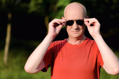 Kıdemli adam yeşil arka planda dururken güneş gözlüğünü ayarlıyor..