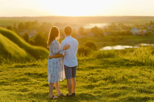 Una joven hermosa embarazada en un vestido largo y su marido de pie juntos en la colina al atardecer —  Fotos de Stock