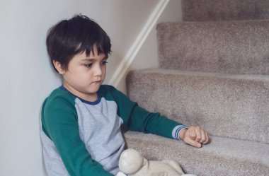 Üzgün Asyalı çocuk pijamalı halı merdivenlerinde oturuyor, üzgün yüzüyle yere bakan yalnız çocuk, depresyondaki çocuk merdivenin köşesinde oturuyor..