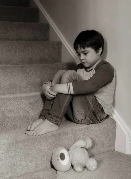 Sad kid sitting on staircase with teddy bear lying down on carpeted in house, Preschool boy looking down with upset face not happy to go back to school, Black and white image, Mental health concept