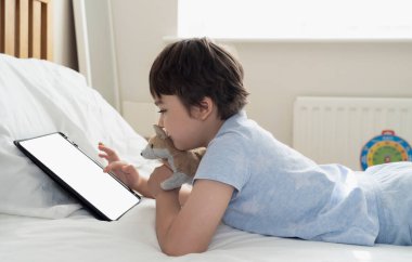 6-7 yaşlarında bir çocuk köpek oyuncağıyla oynuyor ve tabletle çizgi film seyrediyor, Mutlu çocuk sabah yatakta oyun oynuyor, Şirin Çocuk yatak odasında tek başına eğleniyor ve dinleniyor, yeni normal yaşam tarzı. 