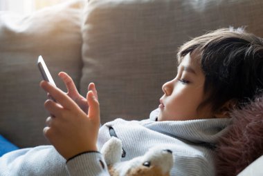 Kırpılmış çocuk kanepeye uzanıp tablette çizgi film seyrediyor, 6-7 yaşında çocuk dokunmatik pedde oyun oynuyor, Şirin Çocuk kendi başına oturma odasında eğleniyor, New normal yaşam tarzı... 