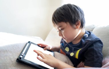 Kırpılmış çocuk kanepede oturmuş tablette çizgi film seyrediyor, 6-7 yaşında çocuk dokunmatik kağıtta oyun oynuyor, Şirin Çocuk kendi başına oturma odasında eğleniyor ve dinleniyor, New normal yaşam tarzı. 