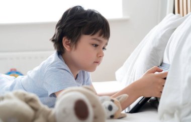 6-7 yaşlarında bir çocuk köpek oyuncağıyla oynuyor ve tabletle çizgi film seyrediyor, Mutlu çocuk sabah yatakta oyun oynuyor, Şirin Çocuk yatak odasında tek başına eğleniyor ve dinleniyor, yeni normal yaşam tarzı. 