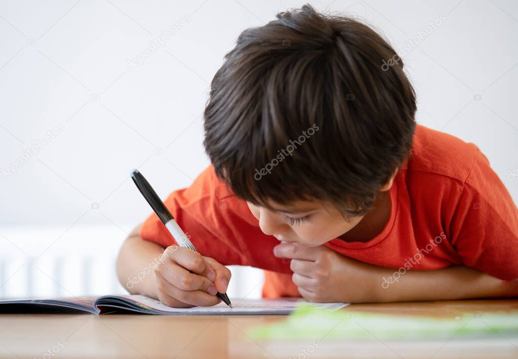Selective focus of school kid boy siting on table doing homework with white background, Happy Child holding black pen,Little boy is writing on white paper at the table,Elementary school and education concept