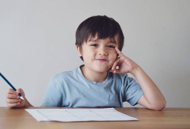 Evde ödev yapan anaokulu çocuğunun portresi. Mutlu Çocuk elinde kalem, gülümseyen yüzüyle kameraya bakıyor, masada yazıyor, ilkokul ve eğitim konsepti.