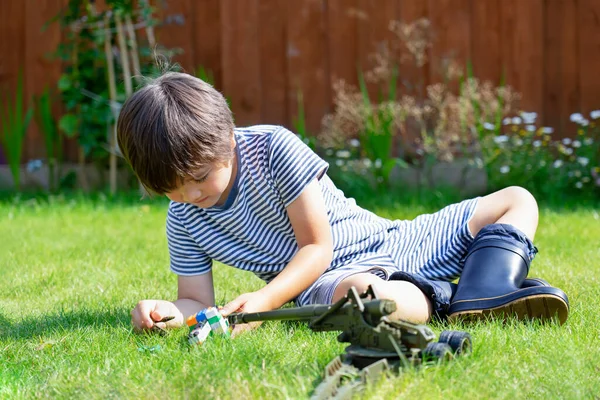 Active boy with excited face lying on grass playing with soldiers and tank toys in the garden, Kid playing wars and peace on his own in hot sunny day, Children imagination and development concept