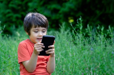 Cep telefonunda mutlu yüz oyunu oynayan açık hava portresi çocuğu, telefonda çizgi film izlerken eğlenen sevimli çocuk, gülen yüz oyunları oynayan çocuk. Network Teknoloji Konsepti