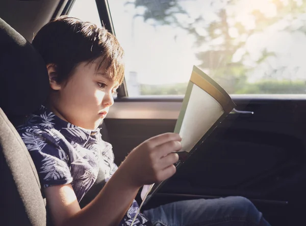 Kid siting on car seat and reading a book, Little boy sitting in the car in child safety seat, Portrait of toddler entertaining himserf on a road trip. Concept of safety taveling by car with children