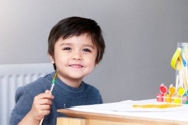 Mutlu çocuk kağıt üzerinde sulu boya çizerken, elinde sanat fırçası tutarken resim çizerken ve evde resim çizerken, soğuk havada çocuklar için kapalı aktiviteler yaparken, çocuk sanatı konsepti yaparken eğleniyor.