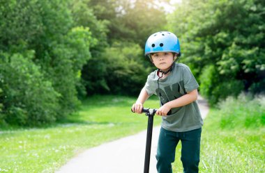 Küçük çocuk güneşli bir yaz günü yeşil parkta scooter sürmeyi öğreniyor, kasklı bir çocuk emniyet kaskı takıyor, çocuk dışarıda oynuyor, çocuklar için aktif eğlence ve açık hava sporları yapıyor..