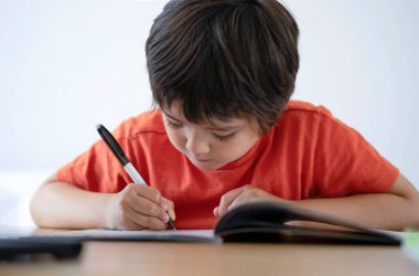 Anaokulu çocuğunun portresi masada oturan beyaz arka planda ev ödevi yapan mutlu çocuk elinde tükenmez kalem olan çocuk masada beyaz kağıda yazı yazıyor ilkokul ve eğitim konsepti.