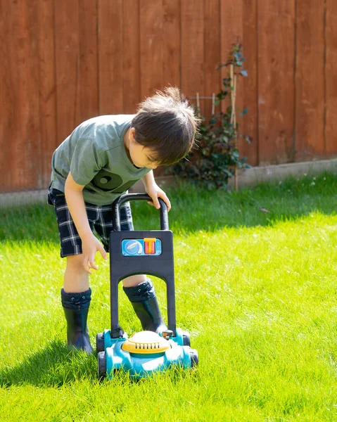 High key light portrait of active kid pushing a toy lawn mower in back yard in summer, Happy child playing with plastic mower toy in the garden in bright light sunny day