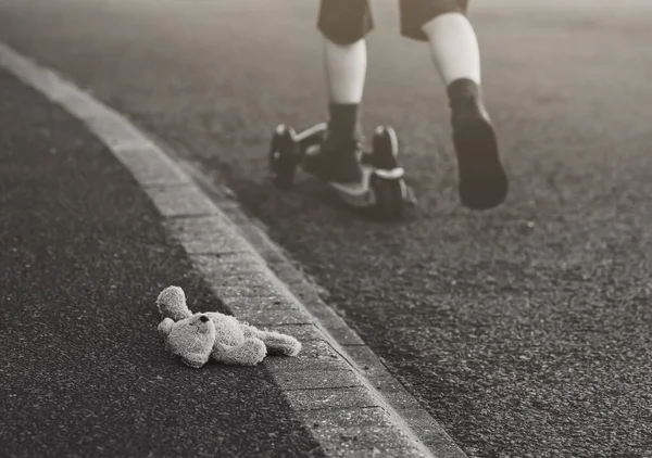 Black and white photo of Teddy bear laying  on the street with blurry background of kid legs run away with scooter, Toy bear was left lying on the street,missing children or kidnap school kids concept