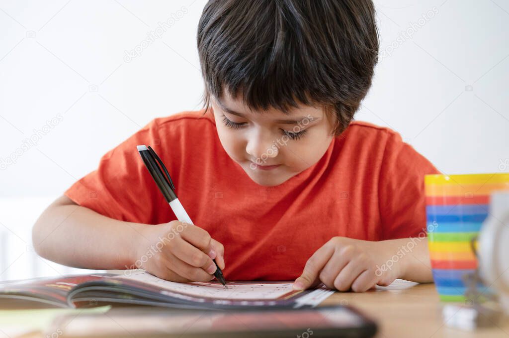 Portrait of school kid boy siting on table doing homework with white background, Happy Child holding black pen,Little boy is writing on white paper at the table,Elementary school and education concept