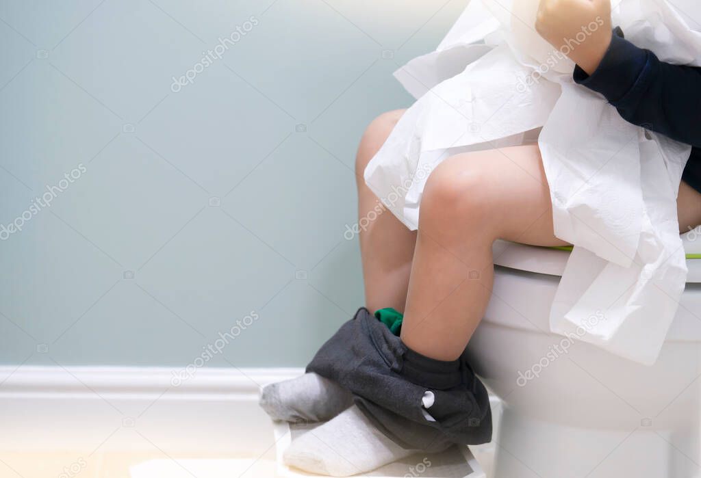 School kid sitting on toilet and playing with toilet rolls, Low view on his legs hanging with grey trousers with fluffy socks, Child tearing the tissue, copy space, Training child or Health care concept.