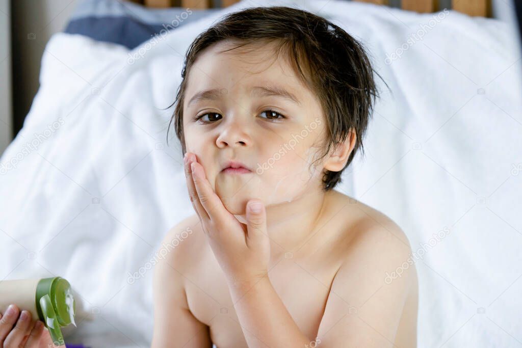 Portrait of cute little boy applying body lotion cream on his cheek after shower,Kid using cream for skincare protection,Child moisturize his dry skin, Children problem,hygiene,clean and fresh concept