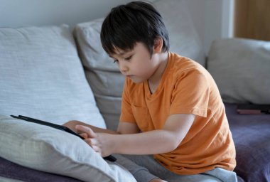 Kırpılmış çocuk kanepede oturmuş tablette çizgi film seyrediyor, 6-7 yaşında çocuk dokunmatik kağıtta oyun oynuyor, Şirin Çocuk kendi başına oturma odasında eğleniyor ve dinleniyor, New normal yaşam tarzı. 