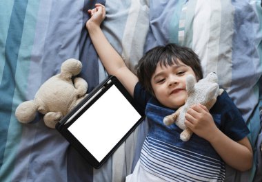 Çocuk yatakta yumuşak oyuncaklarla oynuyor ve gülümseyen yüzüyle Cameara 'ya bakıyor, çocuk tabletle yatakta yatıyor, Top view Çocukları yatak odasında dijital tablet maketi hazırlıyor. Yeni normal yaşam eğitimi 