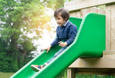 Sevimli küçük bir çocuk oyun parkında retro tonunda kayıyor, sevimli bir çocuk kaydırak evinde oturuyor ve şaşırmış bir şekilde aşağıya bakıyor, güneşli sonbaharda açık havada oynayan aktif bir çocuk.