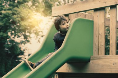 Sevimli küçük bir çocuk oyun parkında retro tonunda kayıyor, sevimli bir çocuk kaydırak evinde oturuyor ve şaşırmış bir şekilde aşağıya bakıyor, güneşli sonbaharda açık havada oynayan aktif bir çocuk.