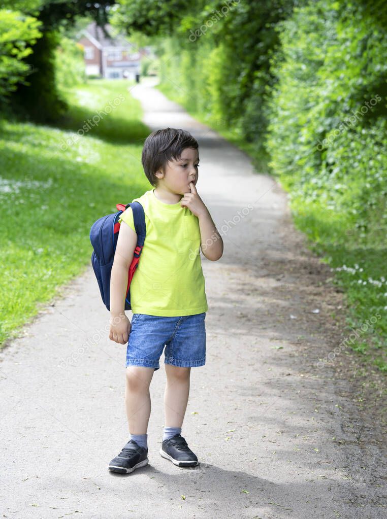 Kid with standing alone and puting finger on his mouth with bored face, Unhappy kid get bored waiting for school bus, School boy carrying backpack get ready for school summer camp
