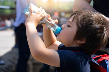 Küçük tatlı çocuk yazın güneşli bir günde berrak cam şişeden soğuk su içiyor, çocuk soda ya da meşrubat içiyor sokakta kalabalığın arasında oturuyor.