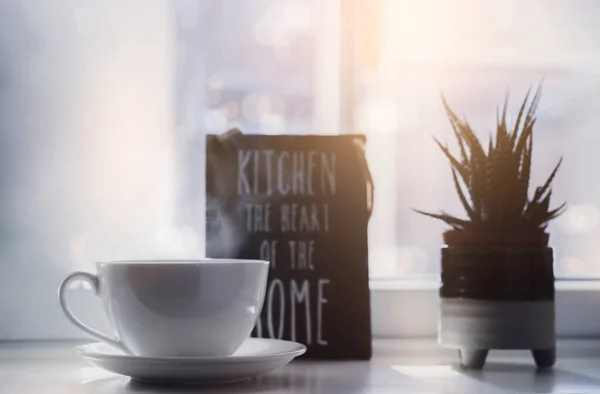 High key light white tea cup with blurry pot of cactus next to window, Hot  cup of coffee  with steam, Cozy and warm sence with bright light shining into kitchen window in the morning