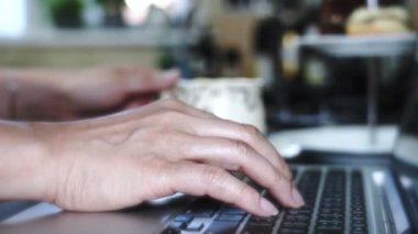 Mutfakta dizüstü bilgisayar kullanan Asyalı bir kadın, klavye defterine yazarken çay ya da kahve içen bir kadın, ev konseptinde online iş yapan yeni normal insanlar.