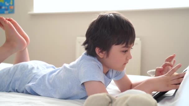 7岁男孩玩狗玩具 在平板电脑上看卡通片 快乐的孩子早上躺在床上玩游戏 可爱的孩子在卧室里玩乐和放松 新的正常生活方式 — 图库视频影像
