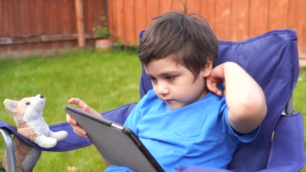 活泼的孩子坐在花园里的椅子上玩游戏或在平板电脑上看卡通片 英俊的男孩在阳光灿烂的夏日在花园里放松 新的正常生活理念 — 图库视频影像