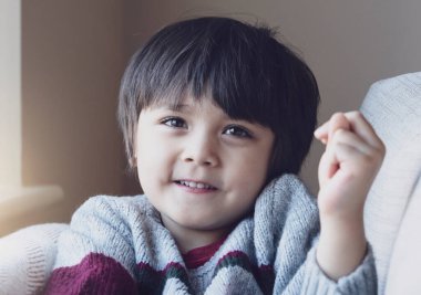 Mutlu çocuk gülümseyen yüzüyle kameraya bakıyor. Sevimli bir çocuğun portresi, gülen yüzüyle kanepede oturuyor, sonbaharda ya da kışın hafta sonlarında evde dinleniyor, sıcak ve rahat bir sahne, pastel tonda..