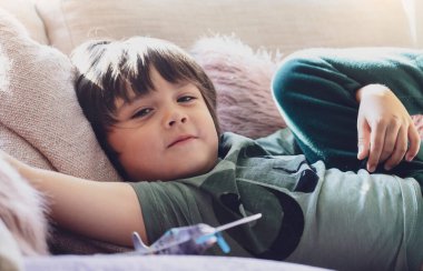 Yüksek anahtarlı, mutlu çocuk koltukta pofuduk yastığa uzanmış seçici odaklı, gülümseyen yüzü hafta sonu evde dinlenen sevimli çocuk, Pastel tonunda sıcak ve samimi bir sahne., 