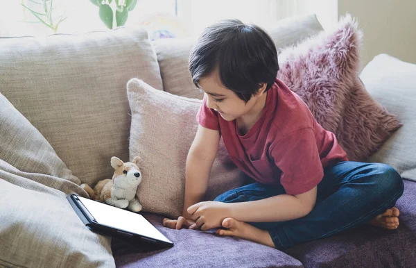 Mixed race kid sitting on sofa watching cartoons on tablet,Portrait 6-7 year old boy playing game on touch pad, Cute Kid having fun and relaxing on his own in living room, New normal lifestyle