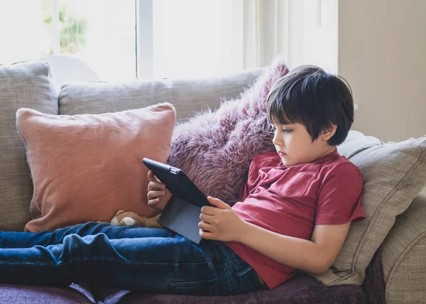 Mixed race kid sitting on sofa watching cartoons on tablet,Portrait 6-7 year old boy playing game on touch pad, Cute Kid having fun and relaxing alone in living room, New normal lifestyle