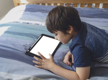 Yeni normal bir hayat, çocuğun ödevi için tablet kullanması, çocuğun yatakta yatıp dijital dokunmatik kâğıtta oyun oynaması, küçük çocuğun tabletle yatması. Evde eğitim, sosyal mesafe, e-öğrenim çevrimiçi eğitim.